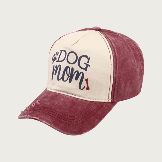 Washed burgundy baseball cap with black 'DOG MOM' text in center (on light beige background), with black & red paw print and red dog bone accent. "Dog" is in all caps serif text, "Mom" is in all caps script text. Rim has 3 small spots of intentional distressing.