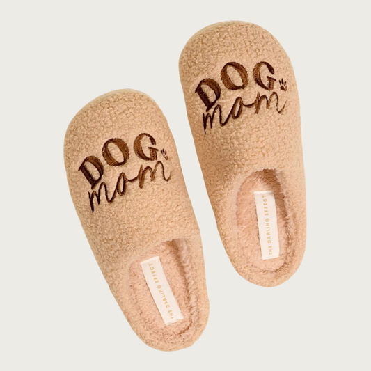 Plush sherpa, tan slippers with "DOG Mom" embroidered in brown on the top of each one. "DOG" is in all caps, and "Mom" is in script text. There is a small brown embroidered paw print on the right, just to the side of the text.