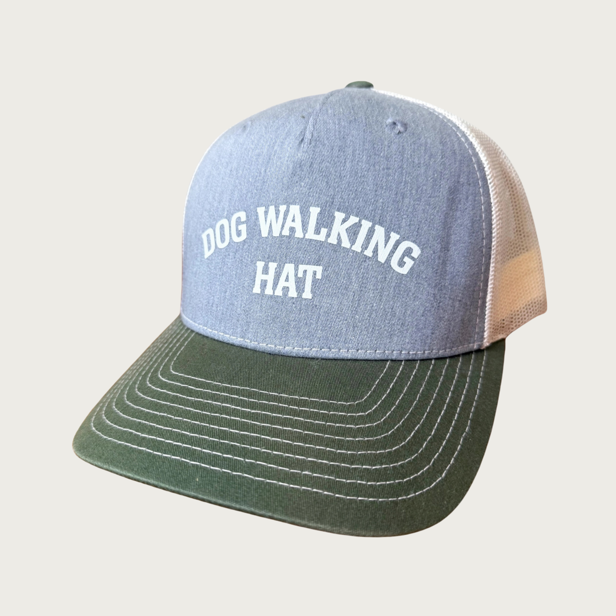 A trucker-style baseball cap with a heathered light blue front panel with white, all caps, arched text reading 'DOG WALKING HAT,' and an olive green flat brim with white contrast stitching. The side & back panels are a warm beige mesh.