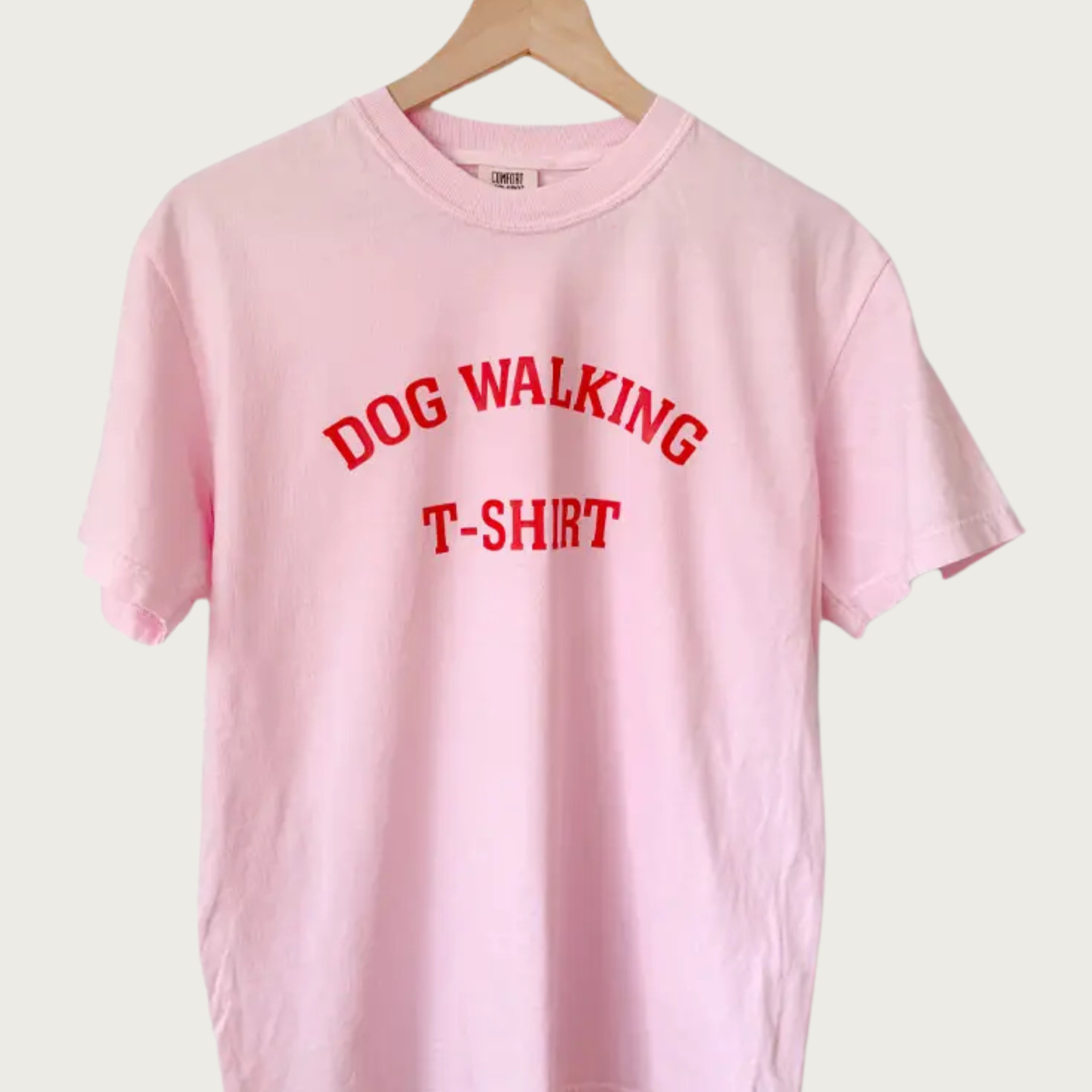 A light pink short-sleeve crewneck t-shirt hanging on a wooden hanger against a light beige background. The shirt features the words 'DOG WALKING T-SHIRT' in arched all caps, red block letters.
