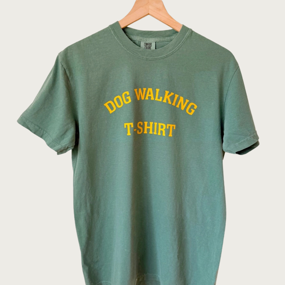 A deep sage green short-sleeved t-shirt hanging on a wooden hanger against a light beige background. The shirt features the words 'DOG WALKING T-SHIRT' in yellow, all caps arched text.