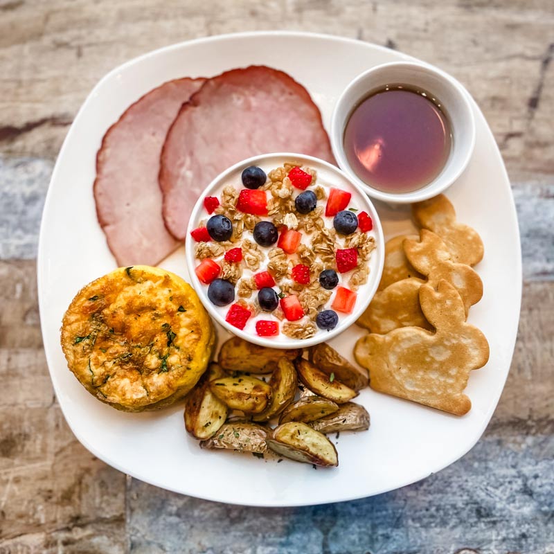 Close-up view of the Easter Brunch meal, displayed on a square, white plate. There are 2 slices of ham, a muffin sized quiche, stack of small roasted potato wedges, 3 bunny-shaped mini waffles with a small side of syrup, and a yogurt bowl topped with diced berries & granola.