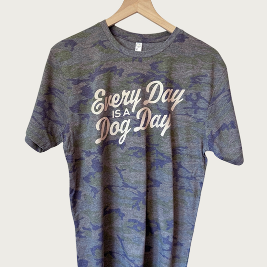 A short-sleeve crewneck t-shirt hanging on a wooden hanger against a light beige background. The shirt features a muted blue, olive green, and gray camouflage print with the text 'Every Day IS A Dog Day' printed in a light beige vintage style script across the chest.