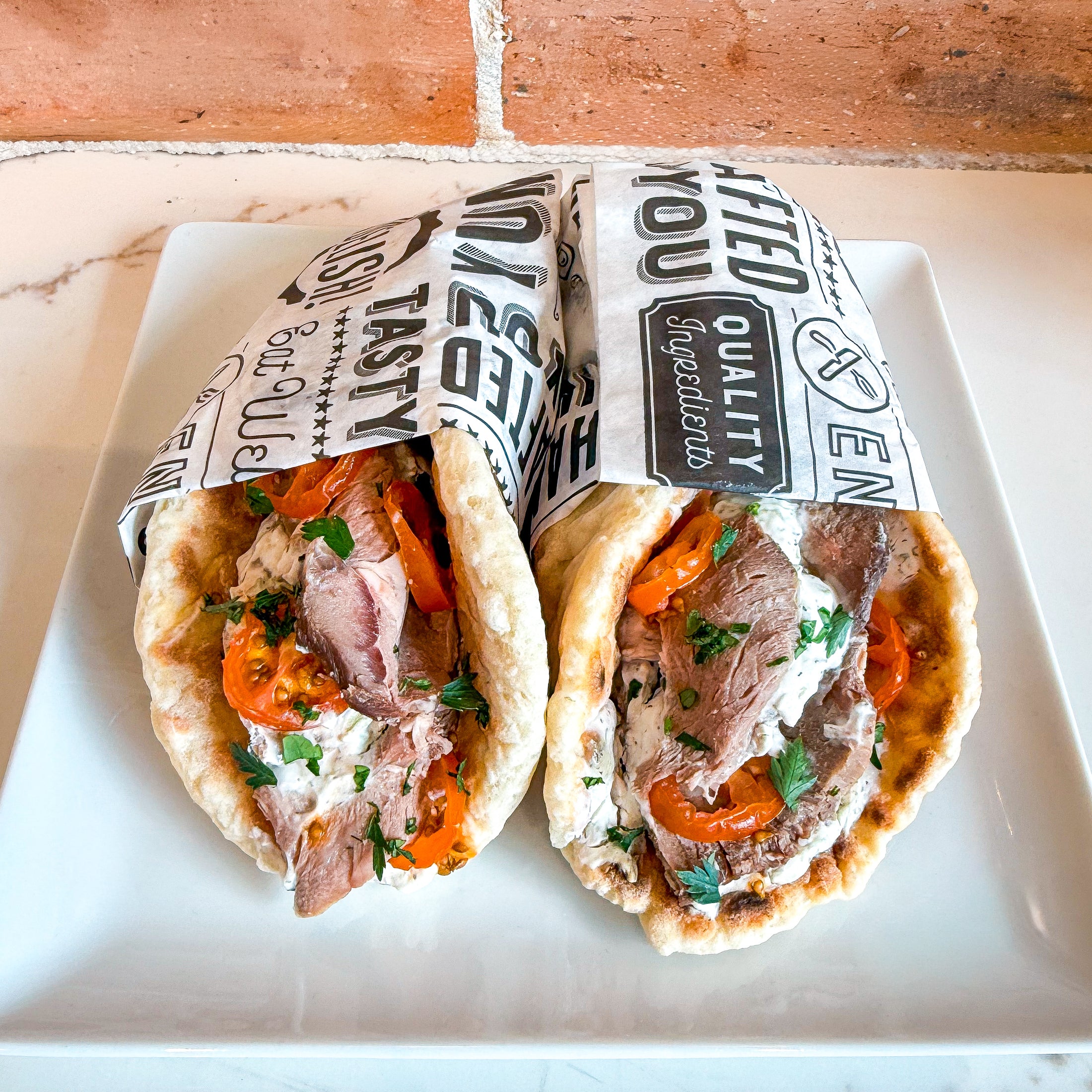 Two stuffed pitas filled with sliced lamb, red tomatoes, creamy sauce and herbs, wrapped in branded paper and served on a white square plate.