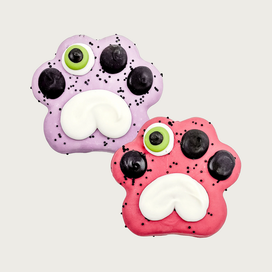 Two paw-shaped dog cookies, one is pale purple and the other a reddish-pink. Design is the same on both: one of the toe pads has a monster eye-white with green and black; the other 3 toe pads are just black. The large, center paw pad is shaped like an upside down, white heart. Both have tiny black sprinkles.