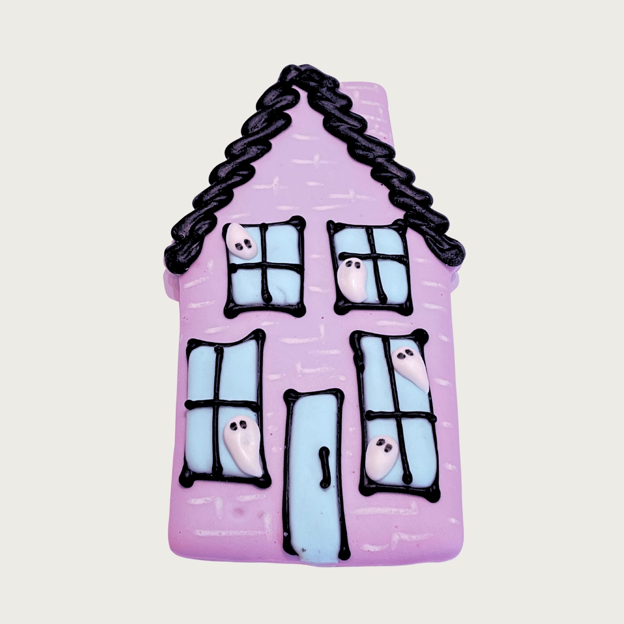Halloween-themed dog cookie, decorated to look like a haunted house. House is lilac with black frosted roofline, with four light blue windows and a door outlined in black. There are white ghosts peeking out of all the windows.