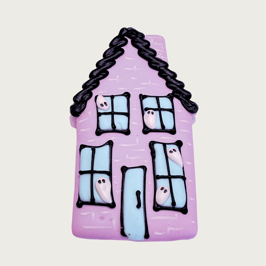 Halloween-themed dog cookie, decorated to look like a haunted house. House is lilac with black frosted roofline, with four light blue windows and a door outlined in black. There are white ghosts peeking out of all the windows.