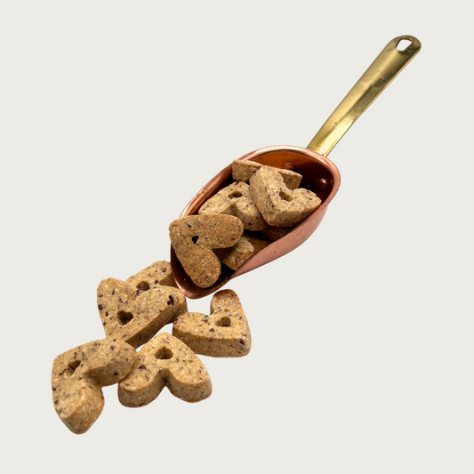 A copper scoop full of heart-shaped dog treats, that are also spilling out in front of it. The individual treats are medium brown in color.