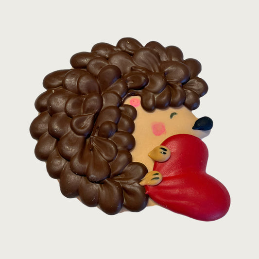 Dog cookie shaped like the side view of a hedgehog. It is holding a red heart with its paws, and has a dark brown back and light brown front, with cute blushing detail on its face.