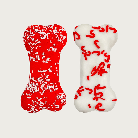 Two bone-shaped, decorated dog cookies, side by side. One is red with white coconut flakes and the other is white with mini red candy cane shaped sprinkles.
