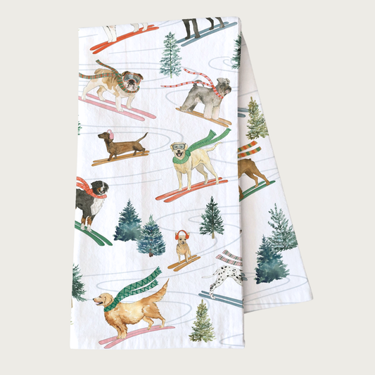 Decorative Christmas-themed tea towel with many different dog breeds on skis, wearing scarves and/or ear muffs, going down a tree-lined ski path. White towel with dog illustrations that looks like they were done in ink & watercolor.