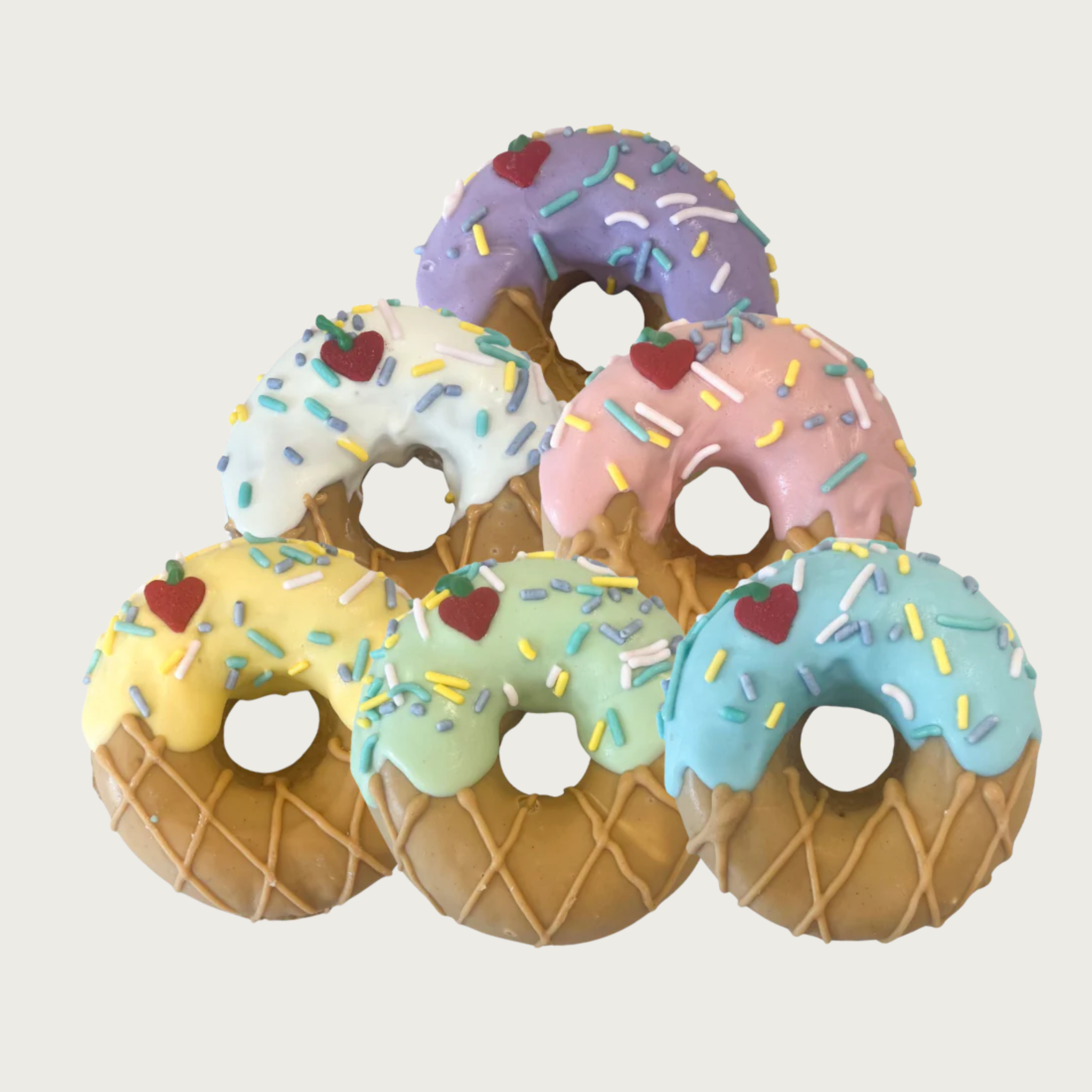 A group of 6 dog cookies, layered in a pyramid shape. Each is decorated to look like an ice cream cone, with tan lower half decorated to look like a waffle cone, and top half with different pastel colored "ice cream" frosting, complete with sprinkles and a heart-shaped cherry-on-top.