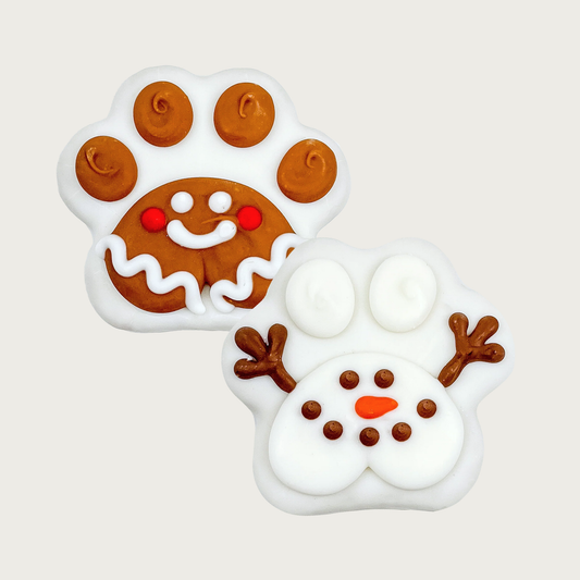 Two Christmas themed paw print shaped dog cookies. Both are covered in a white coating. One has brown toes & brown paw pad, decorated to look like a gingerbread man. The other has a white paw pad decorated to look like a snowman, with 2 white toes and accented with 2 brown sticks for arms.