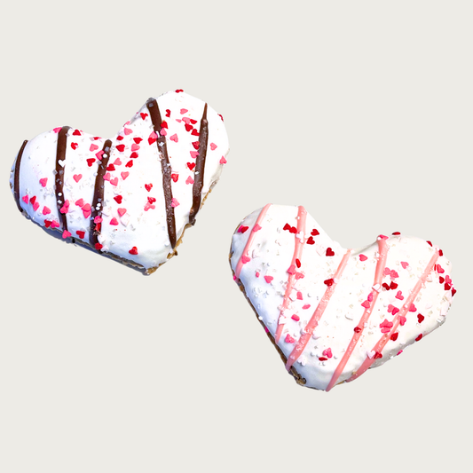 Two Valentine's Day themed, heart-shaped dog cookies with white icing, and tiny red & pink heart sprinkles. One has brown drizzle, the other has pink drizzle.
