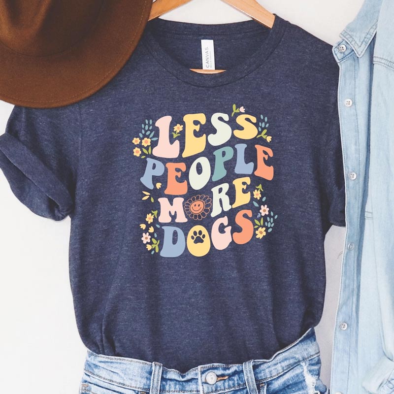 A navy-colored t-shirt with the text 'LESS PEOPLE MORE DOGS' printed in a colorful, 60's style retro, decorative font.