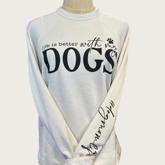An off-white, crew neck sweatshirt. Black text on chest reads, "Life is better with my dogs." The words "with my" are in cursive writing, and the word "dogs" is in extra large print, all caps. There is a paw print icon over the word "my". On the left sleeve, there is text that reads from the elbow down to the wrist, "#dogmomlife"