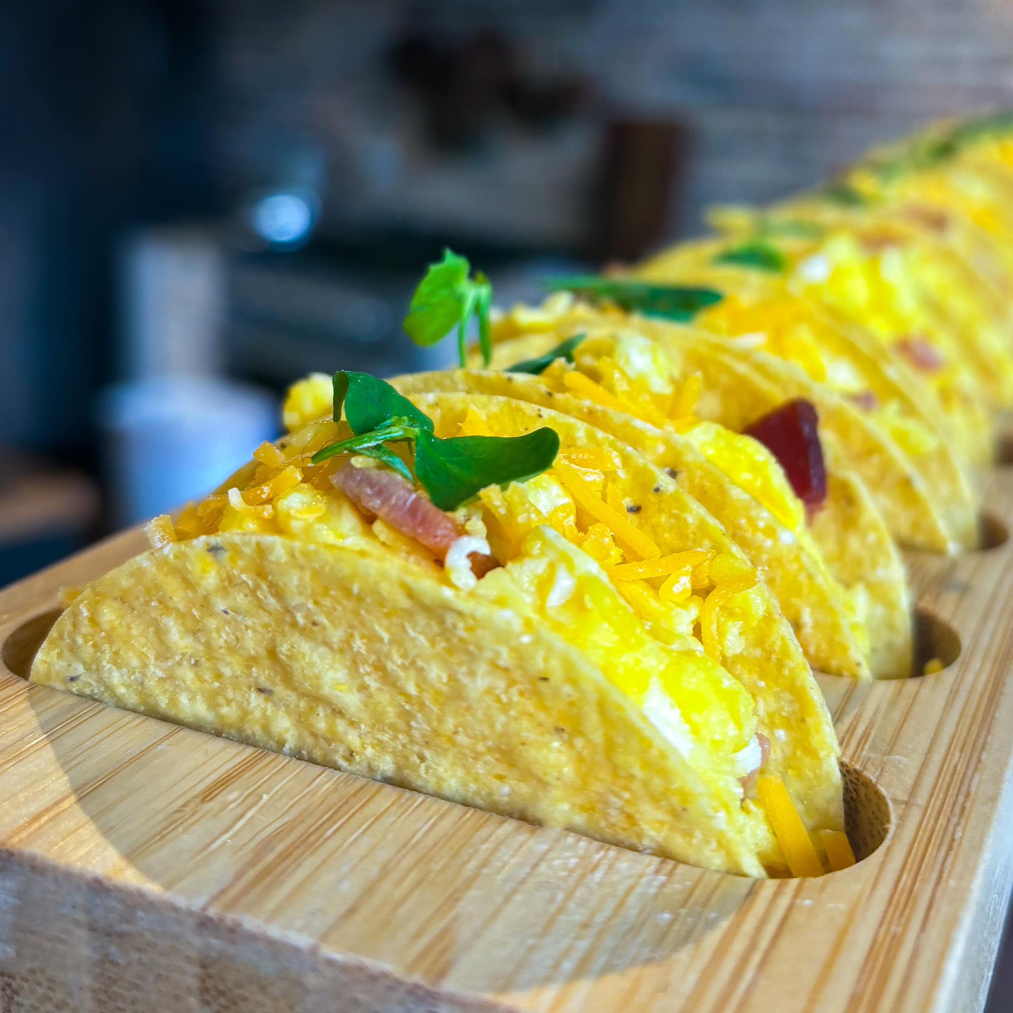Wood tray filled with mini tacos filled with scrambled eggs, shredded cheese, and bacon, and topped with microgreens.