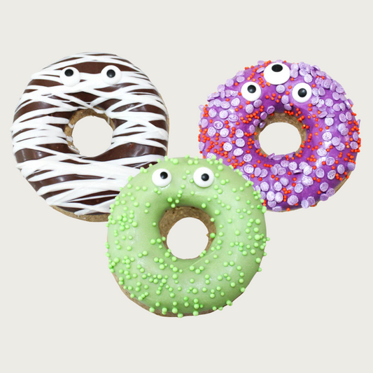 Three thick, donut-shaped dog cookies decorated to look like monsters with googly eyes. One is brown with a vanilla drizzle, the center one is lime green with nonpareils, and the one on the right is purple with red and light purple nonpareils.
