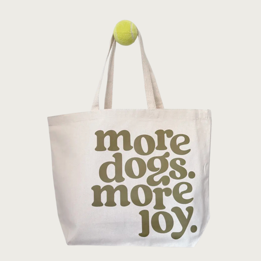 An oversized, cream colored tote bag with two shoulder length handles and an open top. Large, olive green, lower case, right aligned vintage-style text reads "more dogs. more joy.", which fills most of the space on the bag's front.