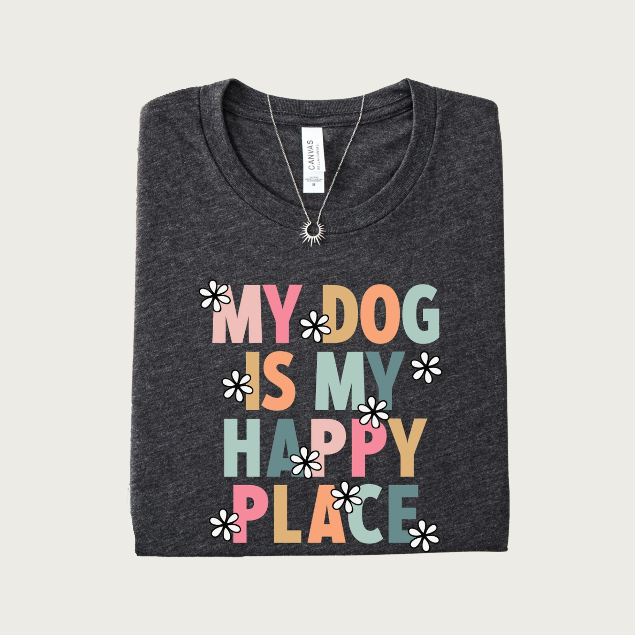 A folded dark heather gray t-shirt with a colorful graphic that reads 'MY DOG IS MY HAPPY PLACE' in shades of pink, orange, and blue-green all caps letters, accented with small black & white daisy flowers.