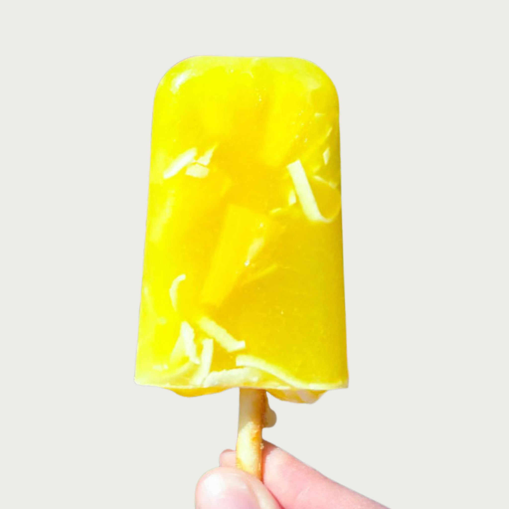A bright yellow popsicle treat for dogs, with visible pineapple chunks and shredded coconut pieces inside, held by its edible No-Hide Chicken Stick.