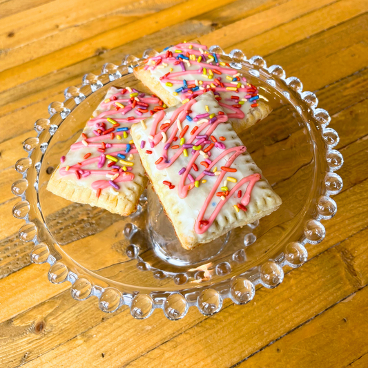 Three rectangular shaped Pup Tart dog pastries with pink icing and sprinkles on a clear glass plate on a wooden surface.