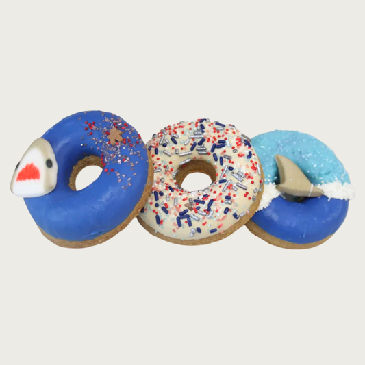 A trio of donut-shaped dog cookies, decorated with a Shark Week theme. One has a shark face popping out of the water (dark blue icing), with a tiny gingerbread man decorette surrounded by red & silver glitter sprinkles. Another is iced in white, with red, blue, & silver colored sprinkles. The last one features a shark fin cutting through the water (dark blue icing with white froth) against a light blue sky.