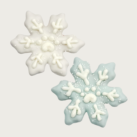 Two snowflake-shaped dog cookies, one white and one light blue, with piped white icing details and white nonpareils.
