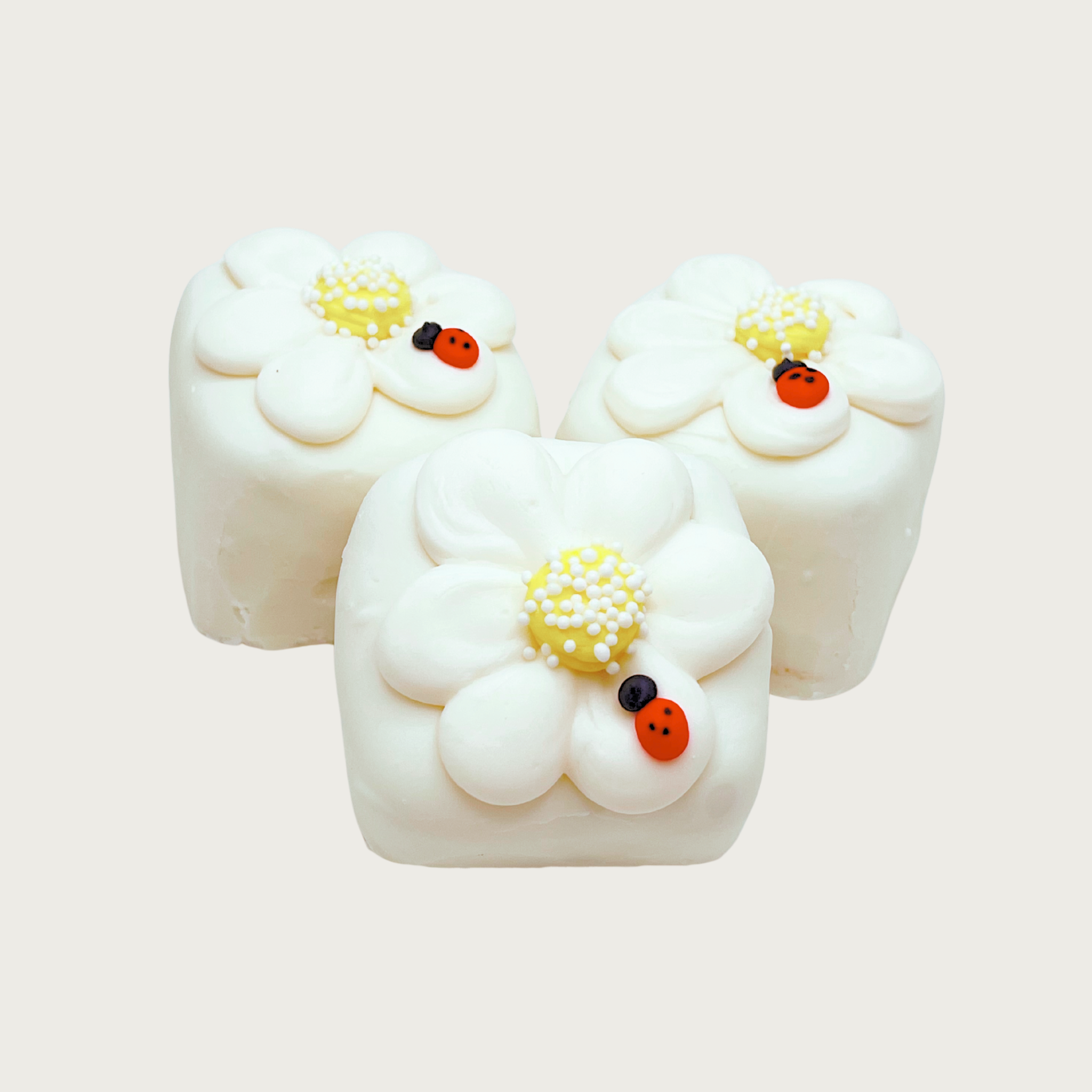 Three square Spring Bon Bon dog treats. They are iced white, and decorated with a large white daisy with yellow center, and small ladybug on bottom right petal.