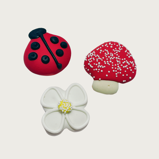 Three small decorated dog cookies. There is a black & red ladybug, mushroom with a red cap and white stem (cap has white nonpareil sprinkles), and a white daisy flower with yellow center, with white nonpareil sprinkles on top of the yellow center.