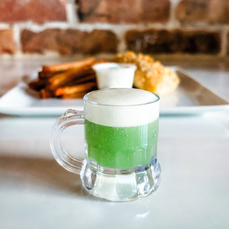 Alternate image of the St. Patrick's Fish & Chips dinner, with the "green beer" in plastic mug as the focal point. The plated dinner is in the background.