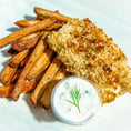 Load image into Gallery viewer, Close-up of the St. Patrick's Fish & Chips dinner, displayed on a white plate. A large portion of golden brown breaded fish is propped on a stack of oven-baked French fries. A small cup of tartar sauce is on the side.
