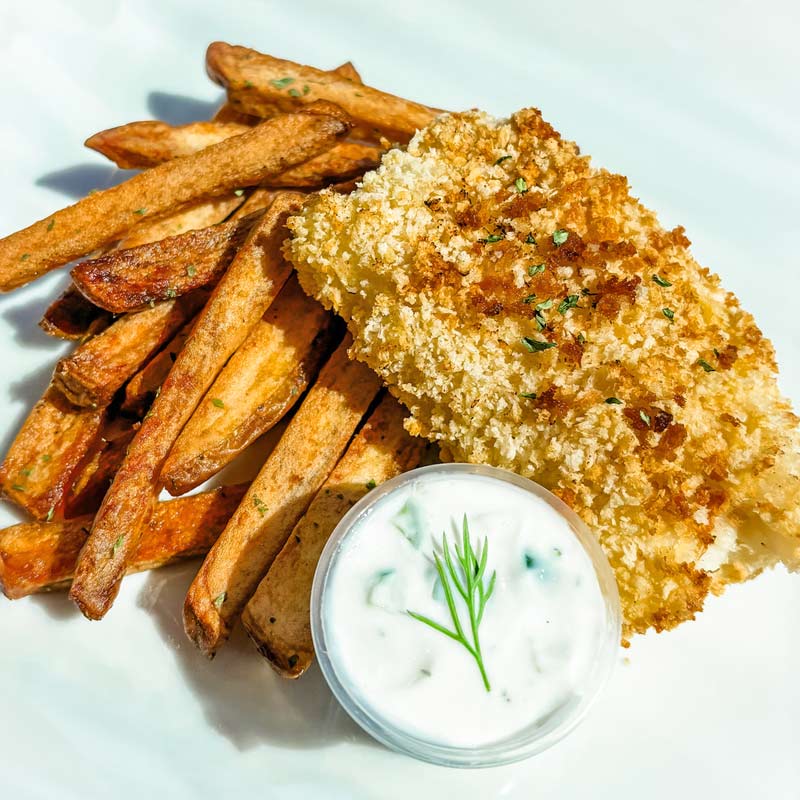 Close-up of the St. Patrick's Fish & Chips dinner, displayed on a white plate. A large portion of golden brown breaded fish is propped on a stack of oven-baked French fries. A small cup of tartar sauce is on the side.