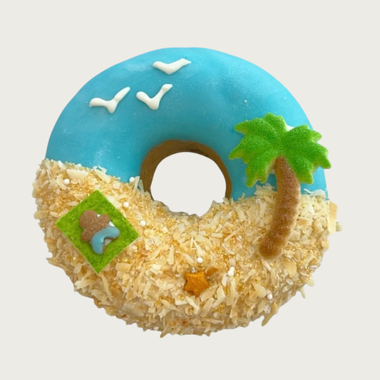 Dog cookie shaped like a donut, decorated like a tropical beach scene with blue icing for the sky, palm tree, and coconut flakes for sand. There are tiny details in the sand such as a sunbather on a towel and little starfish, and iced birds and sunshine in the sky.