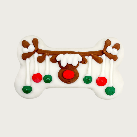 Large bone-shaped dog cookie, covered in white coating. Decorated with Rudolph the Red-Nosed Reindeer, with red & green "ornaments" hanging from his antlers.