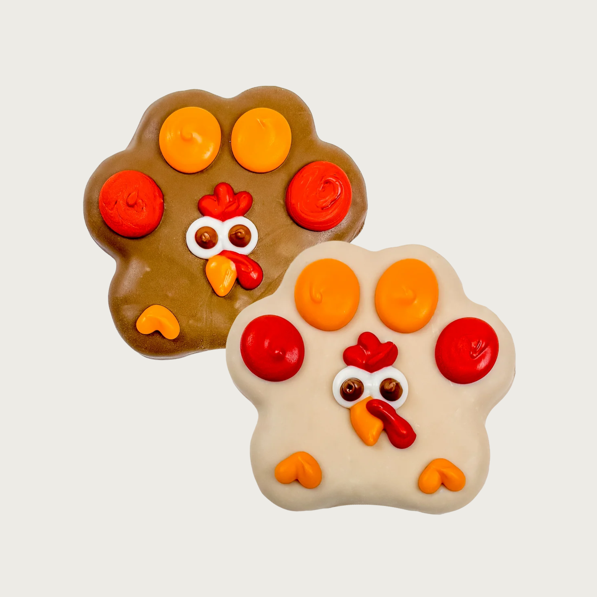 Two decorated, paw print shaped dog cookies, one brown and one beige, both decorated to look like turkeys with white & brown eyes, orange beak, and red wattle. Each "paw pad" is accented with a dollop of either orange or red icing, to look like the turkey's feathers.