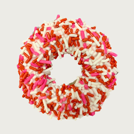 Valentine's themed, donut shaped dog cookie dipped in yogurt coating, and covered with red, hot pink, and ivory colored sprinkles.