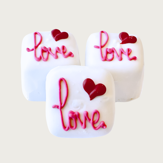 Three square Bon Bon dog treats. They are iced white with "Love" text in hot pink script, and a red heart over the "v."