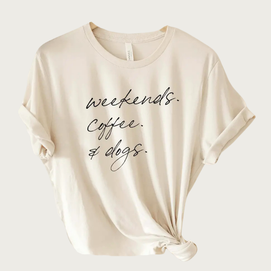 Cream colored t-shirt with crew neck, and 'weekends. Coffee. & dogs.' text in thin, black script font. Each word is on its own separate line, and the entire text block is centered.