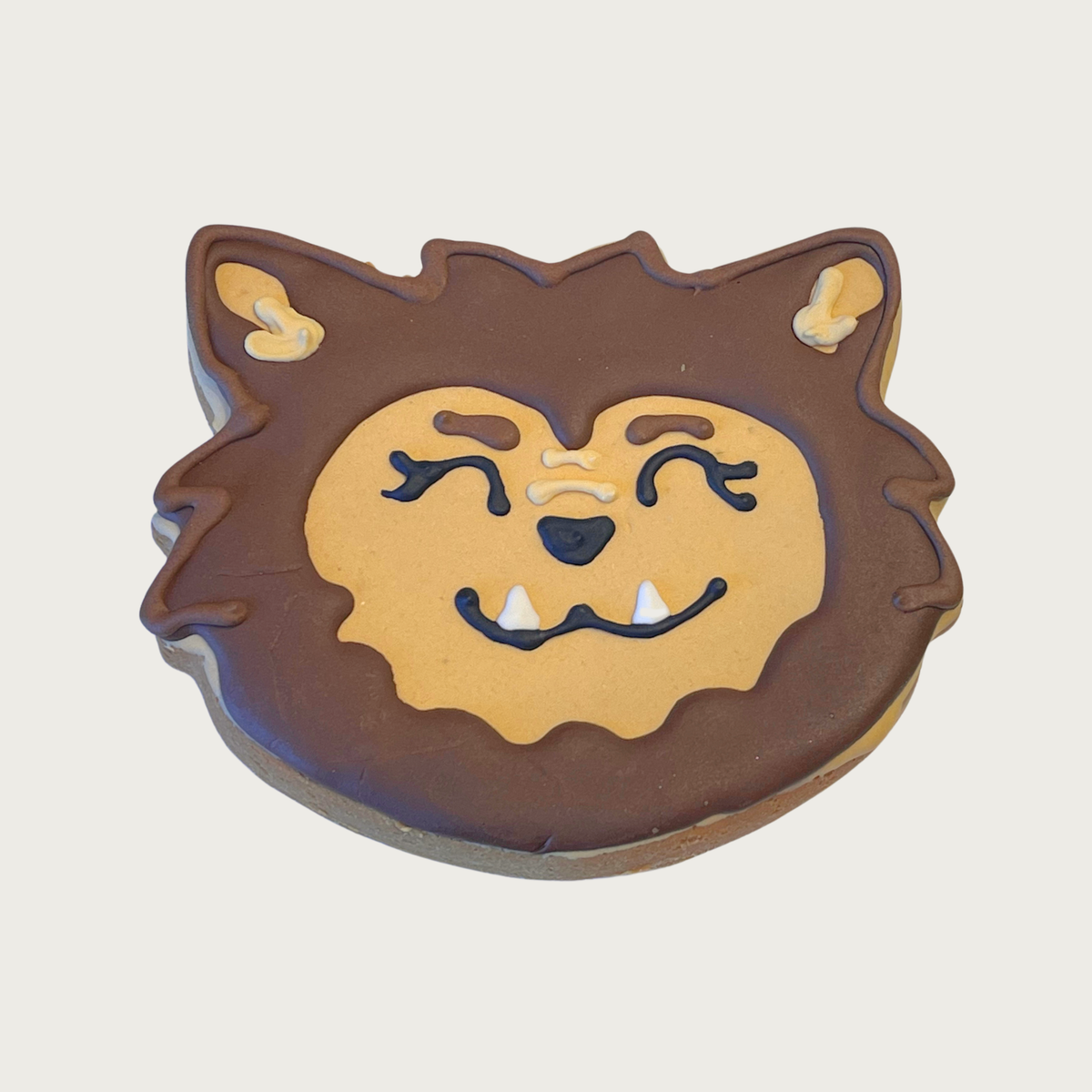 A dog cookie decorated to look like a werewolf's head, featuring brown and beige frosting, black eyes and nose, and white fangs, with a friendly facial expression.