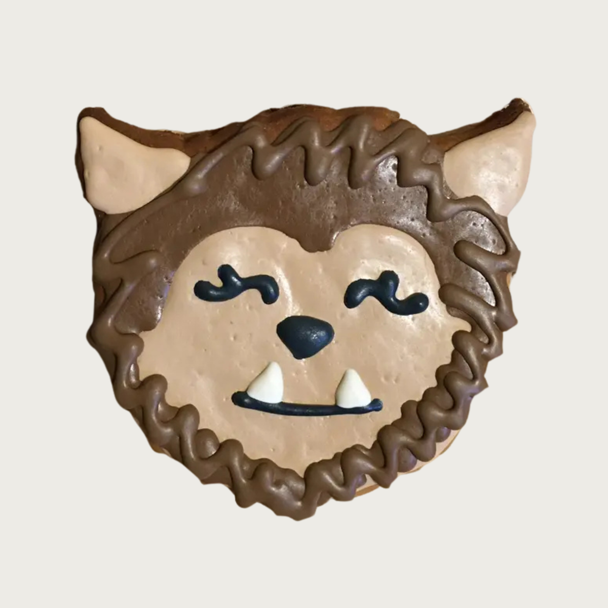A dog cookie decorated to look like a werewolf's head, featuring brown and beige frosting, black eyes and nose, and white fangs, with a friendly facial expression.