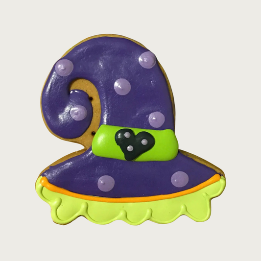 A decorated witch hat dog cookie with purple icing, lavender polka dots, a lime green band with a black heart, and a scalloped lime green edge.