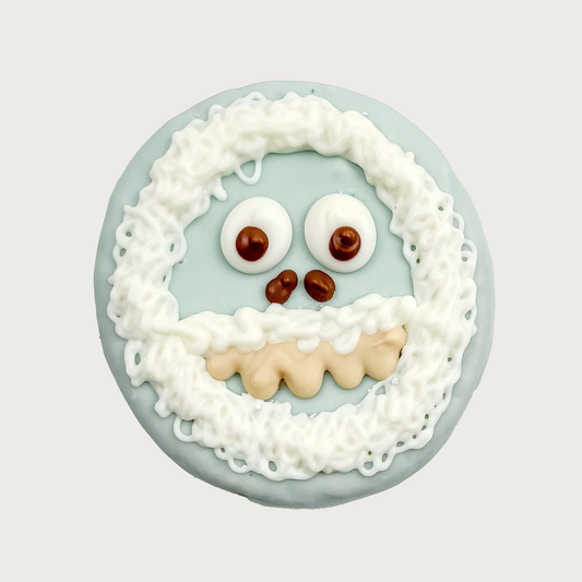 Dog cookie decorated to look like a yeti, or abominable snowman. It's covered in pale blue icing, with white icing in thick drizzles to look like fur around its face and mouth. It has large, googly eyes, small nostrils, and cream-colored teeth.