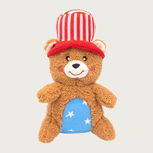 Stuffed plush brown teddy bear dog toy wearing a red and white striped top hat. Its belly is light blue fabric with white stars pattern. Embroidered smiley face expression.