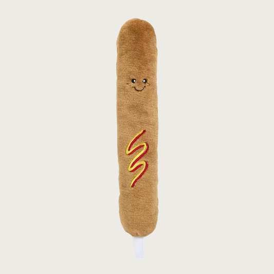 A plush corn dog shaped dog toy with a brown fuzzy body, embroidered smiling face, and red and yellow wavy mustard & ketchup details in the center of its body.