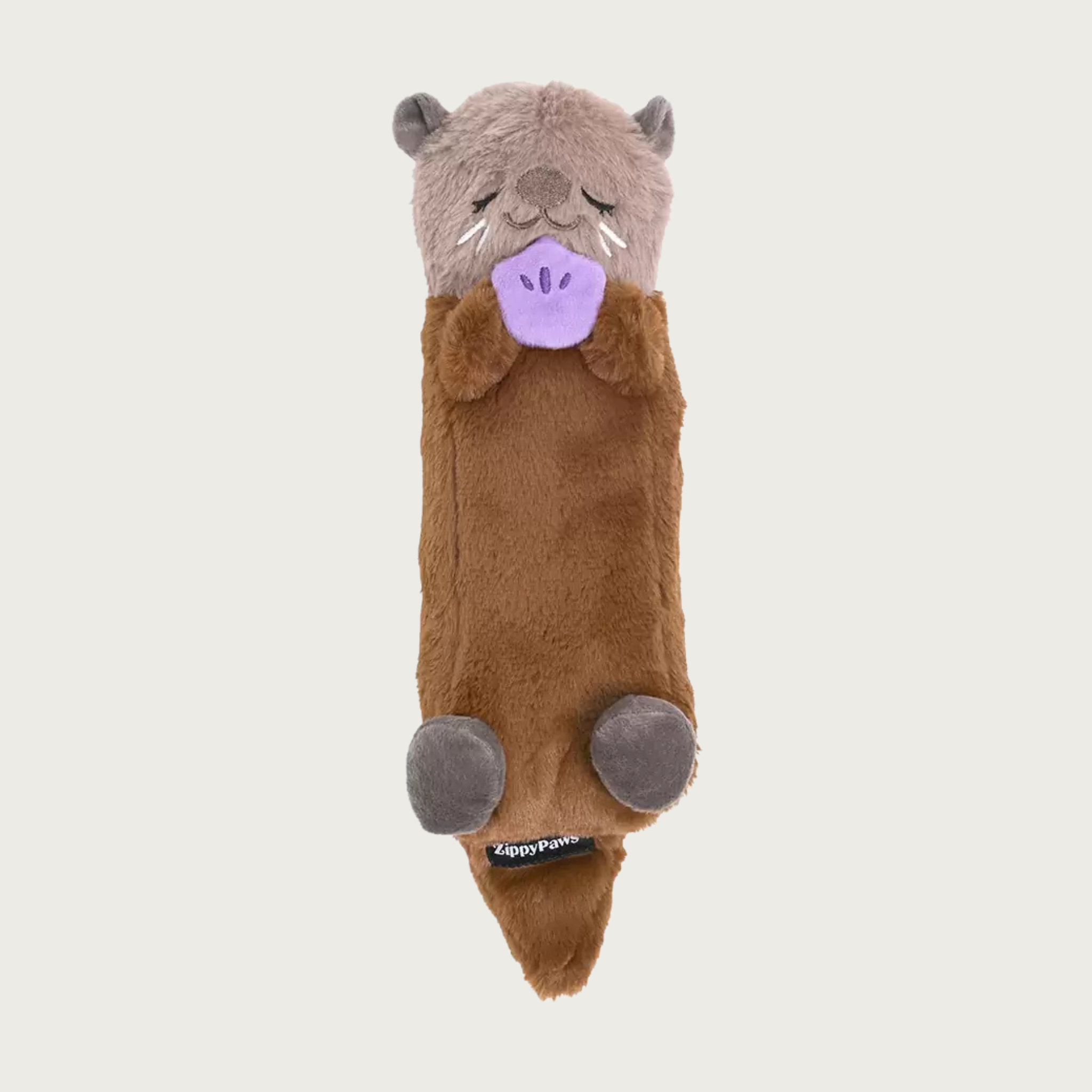 A long, plush sea otter dog toy, laying on its back and holding a purple clam between its front paws. Its body is brown, its head is a lighter brown with a sleepy embroidered facial expression, and it has gray ears & feet.