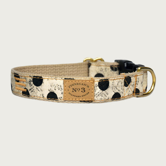 Creamy beige dog collar with large black polka dots, with a subtle, muted black swirl pattern behind the dots. Black plastic buckle and brass D-ring.