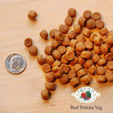 A close-up image of a handful of dry Beef Frittata Veg kibble pieces, next to a dime for size comparison. About 3 of the pieces would fit into the area of the dime. They are small, and about the size and shape of a pencil eraser.
