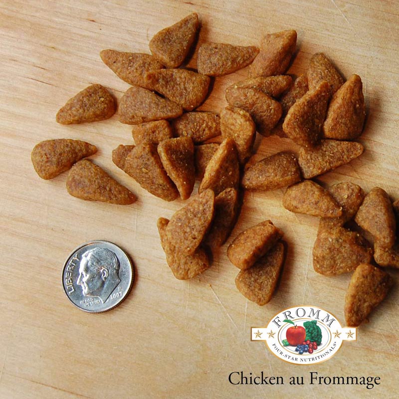 A close-up image of a handful of dry Chicken au Frommage kibble pieces, next to a dime for size comparison. 1-2 of the pieces would fit into the area of the dime. They have a slightly elongated triangle shape to them.