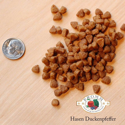 A close-up image of a handful of dry Hasen Duckenpfeffer kibble pieces, next to a dime for size comparison. About 5-6 of the pieces would fit into the area of the dime. They are very small and have a rounded triangle shape.